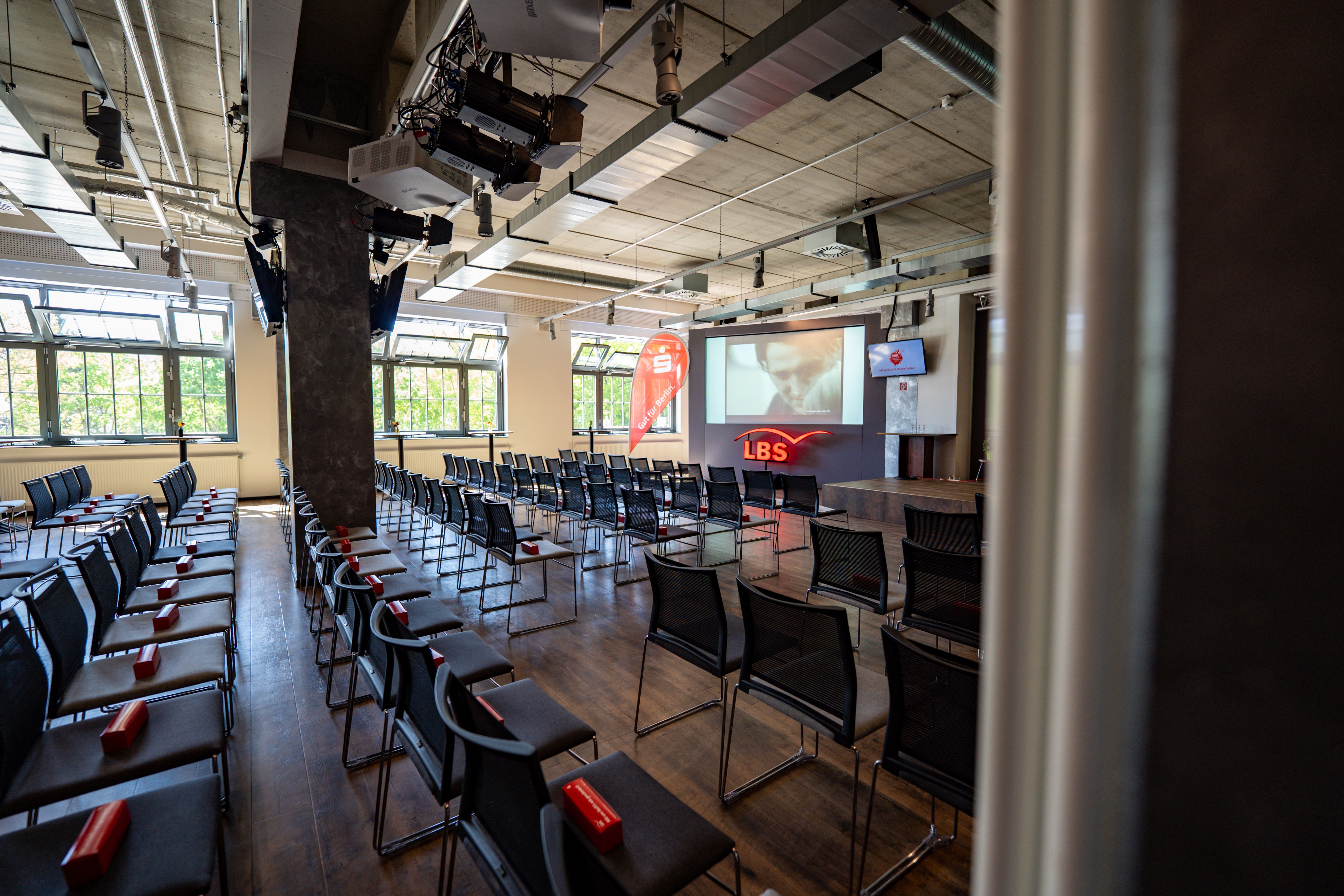 Ein moderner Veranstaltungsraum mit Reihen von Stühlen, einer Präsentationsfläche und großen Fenstern.