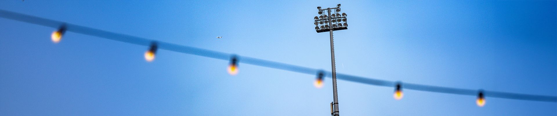 Ein Flutlichtmast vor einem blauen Himmel, im Vordergrund eine Lichterkette mit mehreren Leuchten.