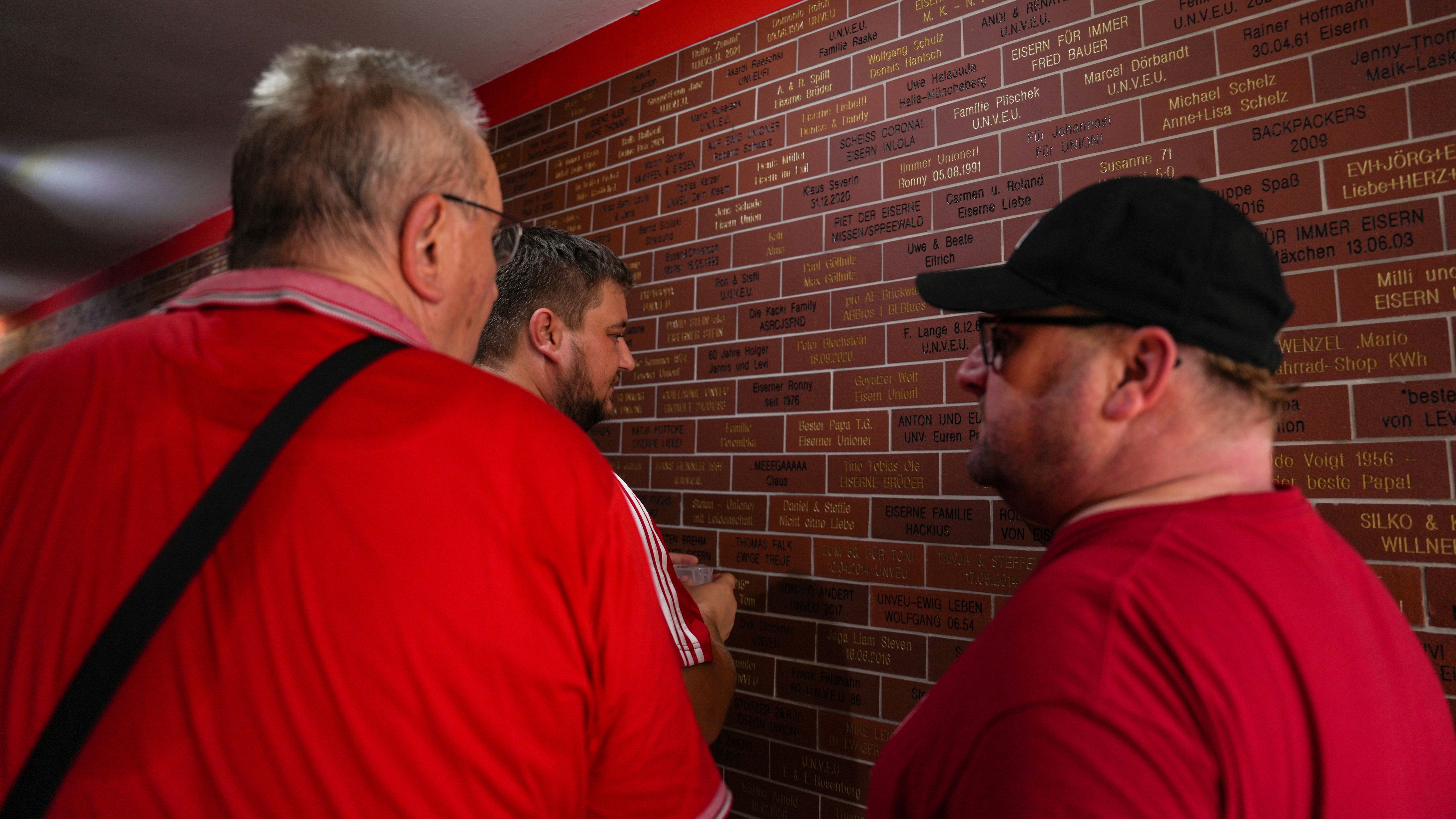 Drei Männer betrachten eine Wand mit vielen braunen Plaketten, die Namen und Daten zeigen.