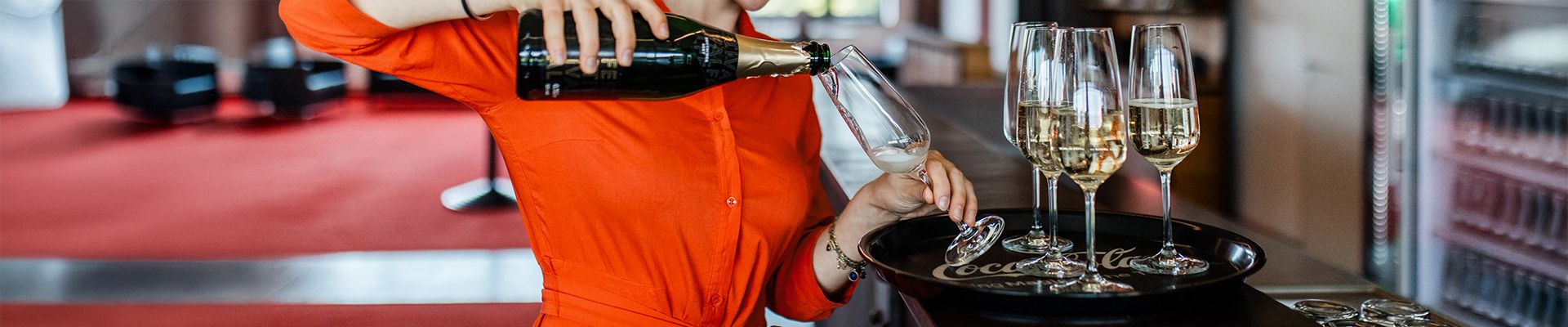 Eine Person in orangefarbenem Oberteil schenkt Sekt in ein Glas auf einem Tablett. Im Hintergrund stehen weitere Gläser.