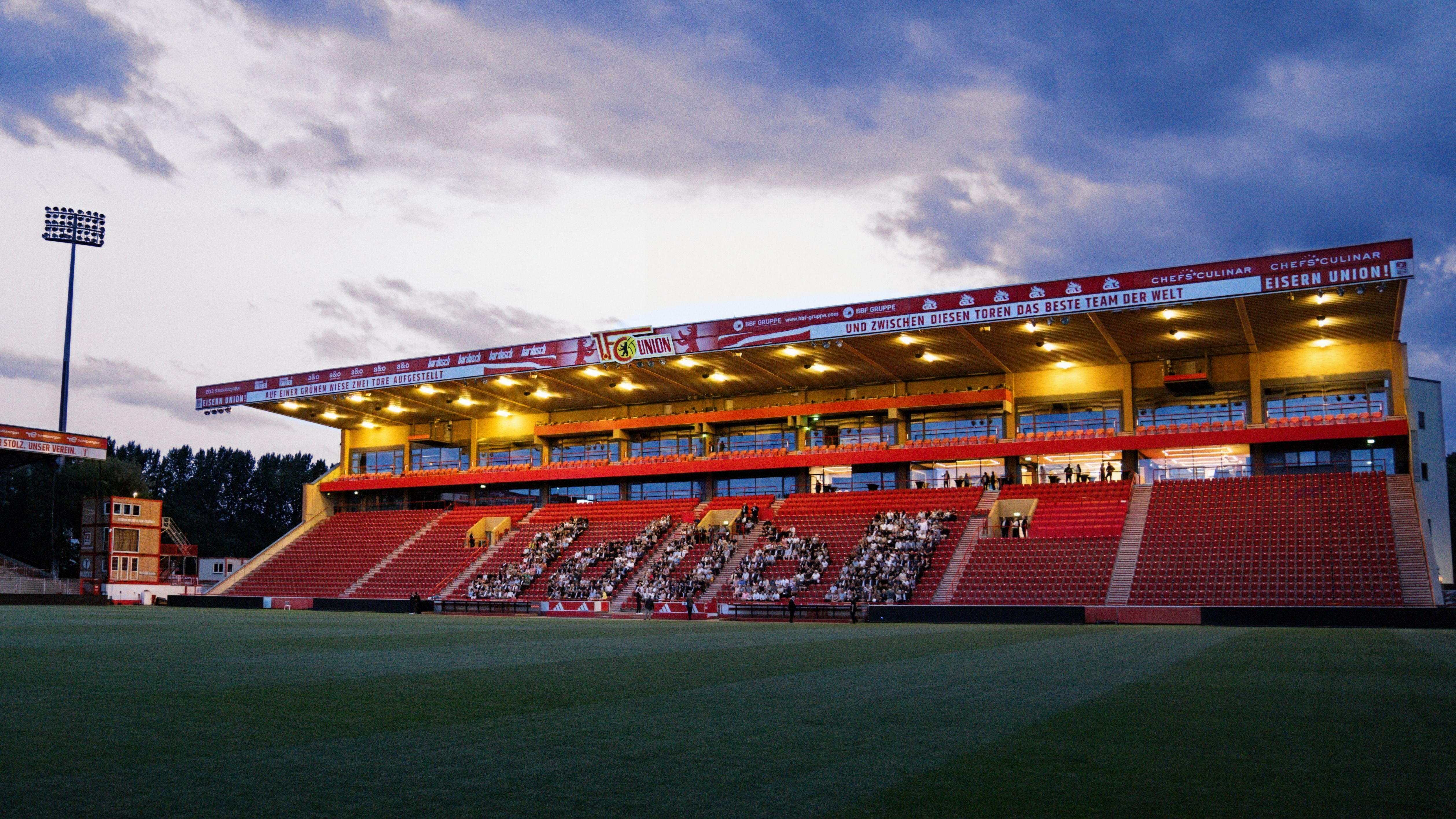 Eine leere Fußballtribüne bei Dämmerung, mit Wolken am Himmel und einem beleuchteten Stadion.