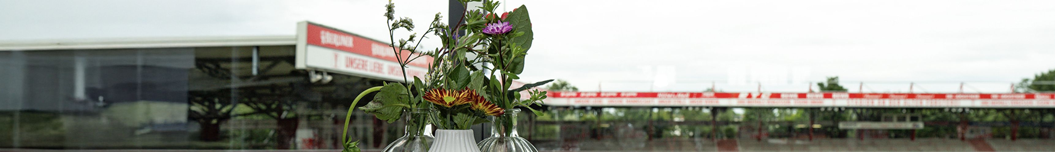 Vase mit bunten Blumen im Vordergrund, im Hintergrund ein Sportstadion mit Tribünen.