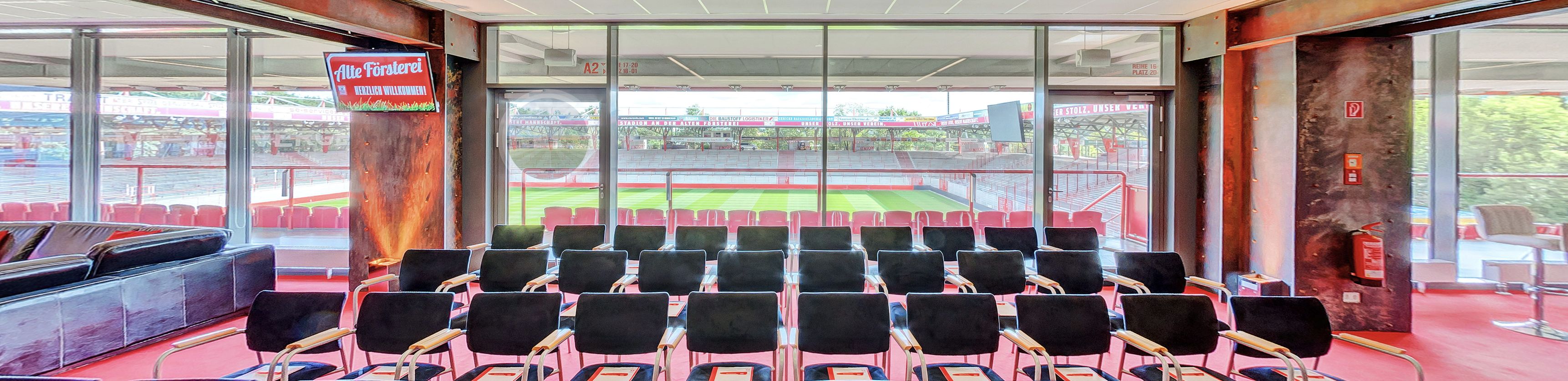 Blick auf ein modernes Stadioninnere mit Sitzreihen, großen Fenstern und einem grünen Spielfeld im Hintergrund.