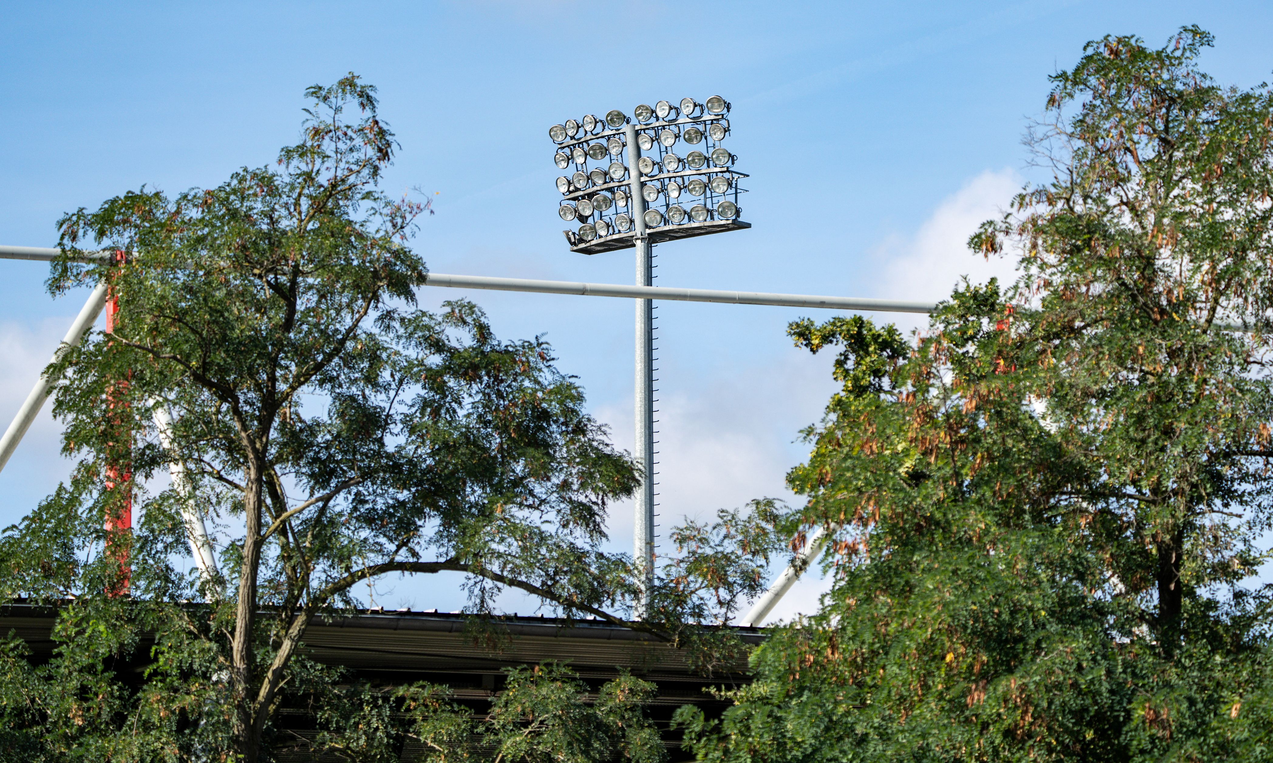 Flutlichtmasten über Bäumen, leicht wolkiger Himmel im Hintergrund.