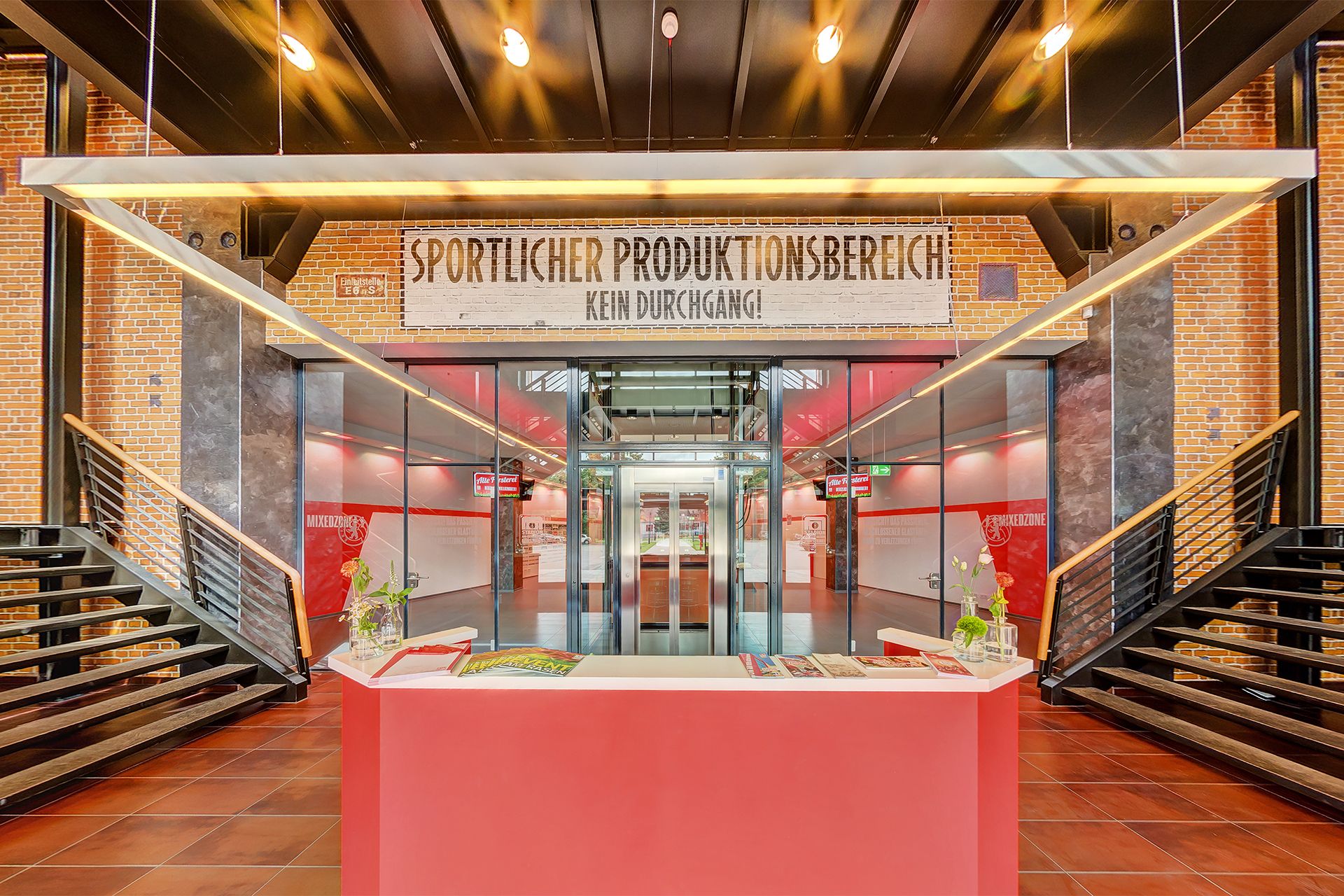 Eingangsbereich mit rotem Empfangstresen, Backsteinwänden und großen Aufzügen, beschriftet mit "Sportlicher Produktionsbereich".