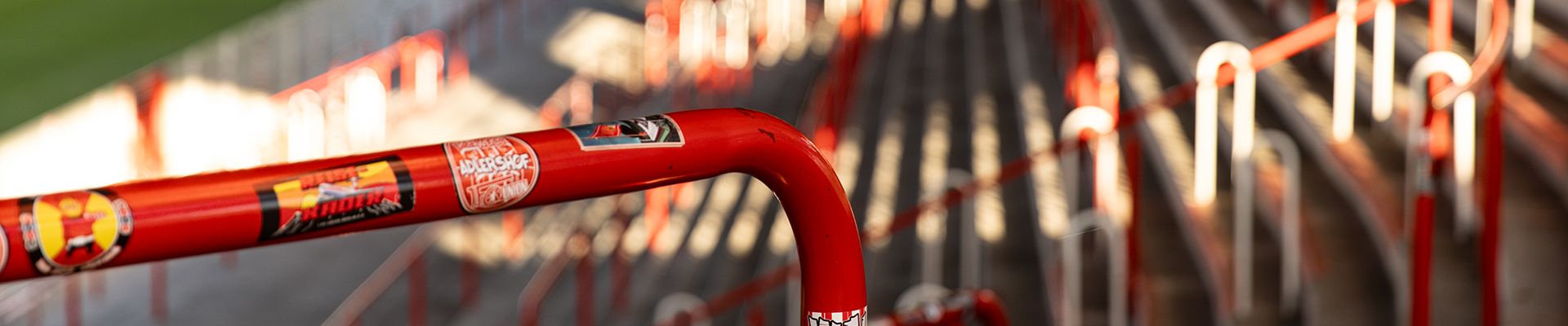 Nahaufnahme eines rot lackierten Handlaufs mit Aufklebern, im Hintergrund verschwommene Stadionstufen.
