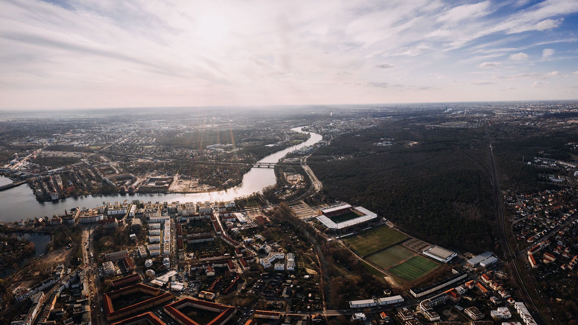 Luftaufnahme einer Stadtlandschaft mit einem Fluss, umgeben von Gebäuden, Wäldern und einem Fußballfeld im Vordergrund.