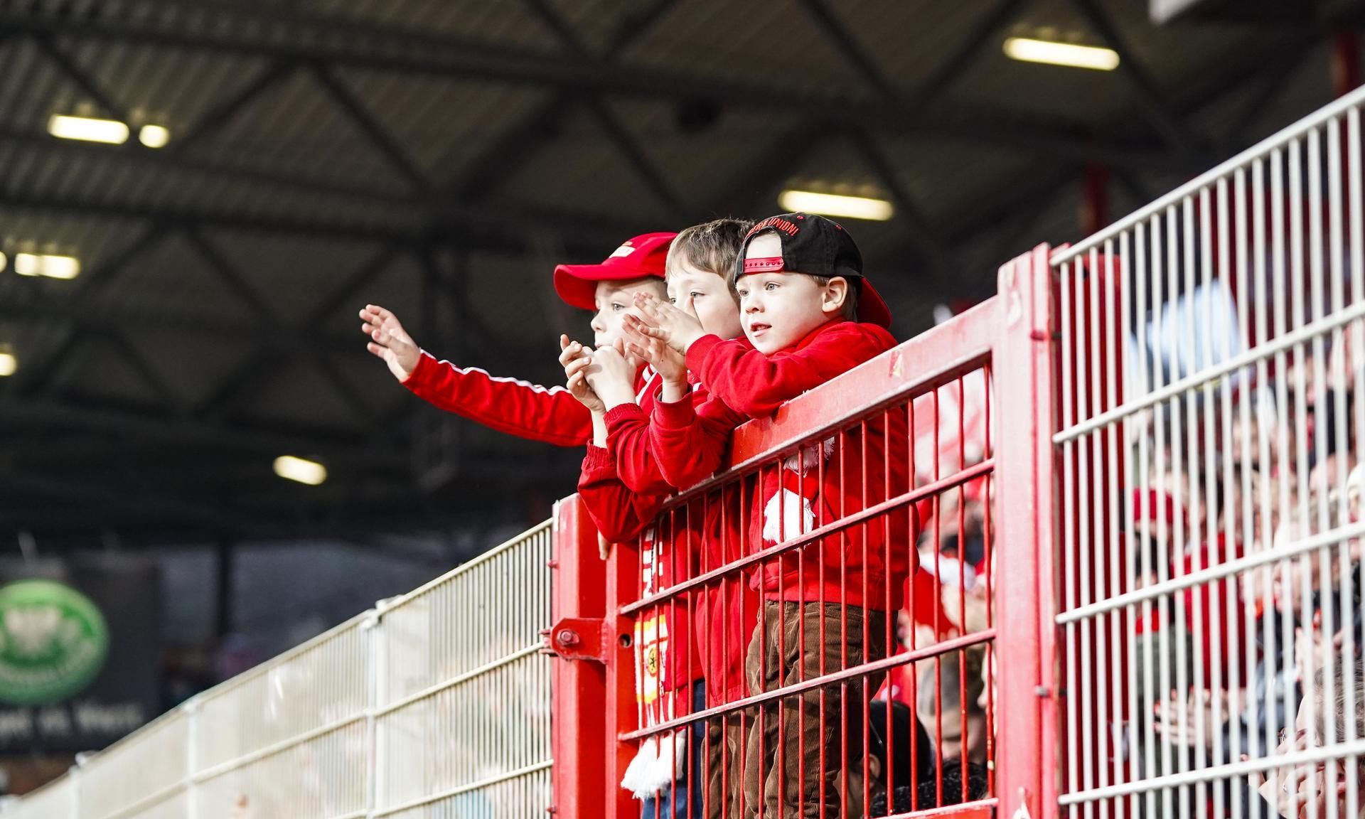 Drei Kinder in roten Trikots stehen an einer Tribünenbarriere und applaudieren begeistert bei einem Sportereignis.